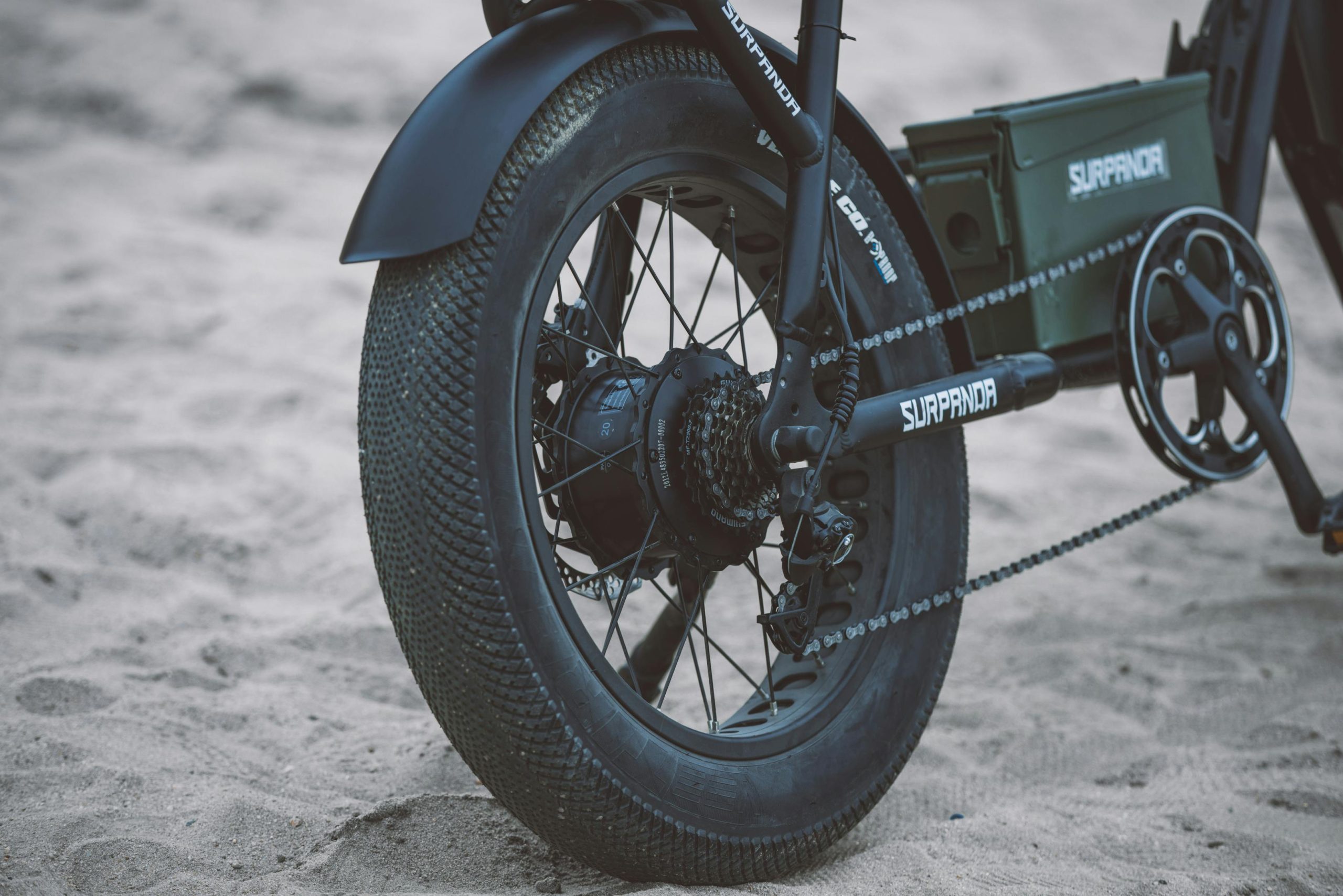 Close-up view of electric bicycle wheel on sandy beach, highlighting the tire and chain.